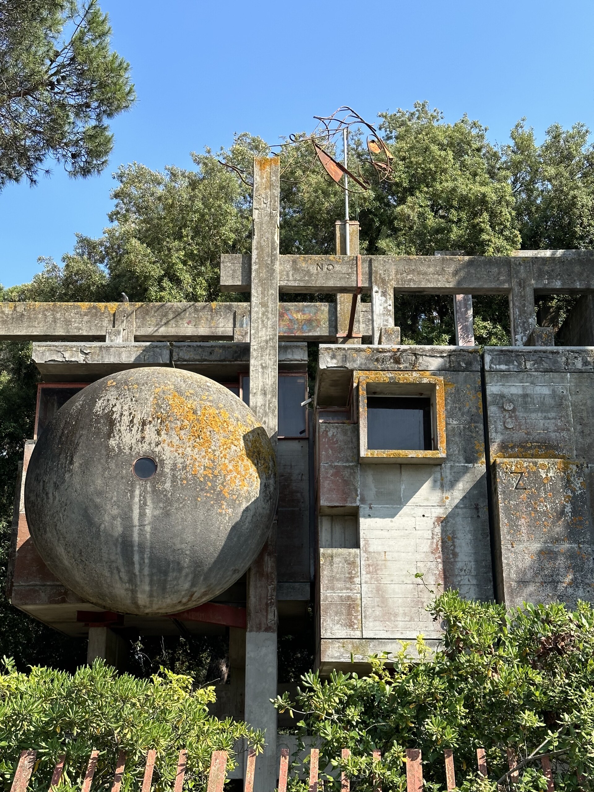Casa Sperimentale - Unique concrete structure with large spherical and rectangular shapes, covered in moss and weathered textures. Set against a backdrop of green trees and blue sky. A metal sculpture resembling a weathervane tops the building.
