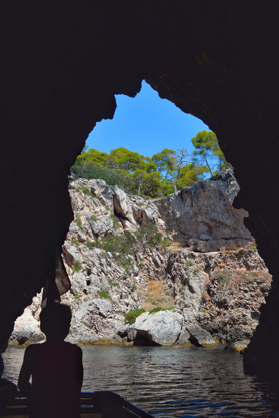 Isole Tremiti - View from inside a dark cave looking out towards a rocky landscape with green trees and a clear blue sky. Silhouette of a person in a boat in the foreground.