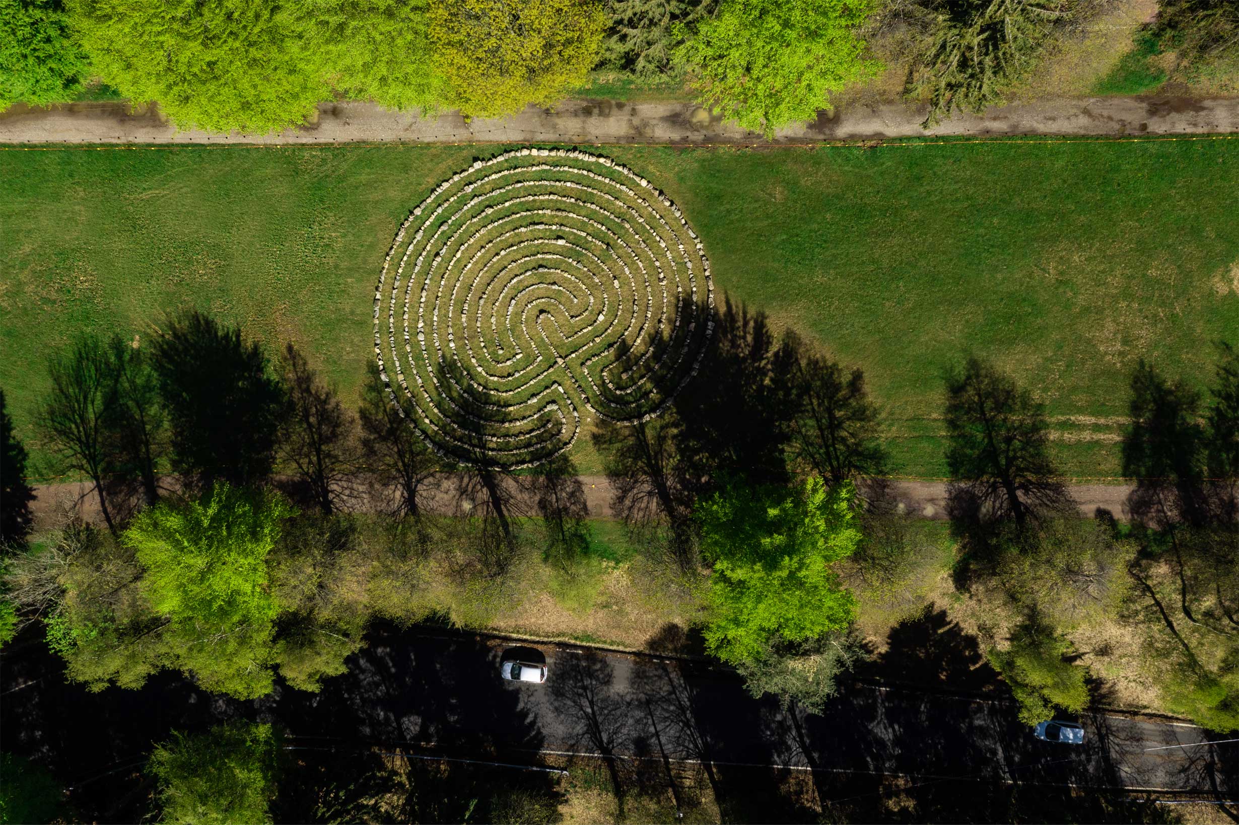 [Vista aerea di un labirinto circolare di pietra] - Vista aerea di un labirinto circolare di pietra su un campo erboso, circondato da alberi che proiettano lunghe ombre. Un sentiero e delle auto parcheggiate sono visibili nella parte inferiore dell'immagine.