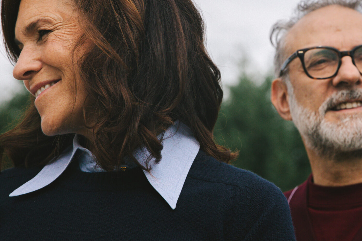 [No Title] - A close-up of two adults outdoors, with a woman in the foreground smiling and a man in glasses behind her, both partially out of frame. The background is blurred greenery.