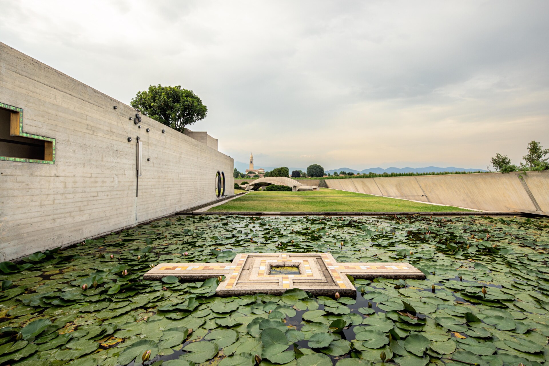 Tomba Brion - Reflecting pool with geometric tile design surrounded by lily pads in a modern courtyard. Concrete walls border the space, featuring an alcove and tree. A grass lawn stretches towards distant trees and mountains under a cloudy sky.
