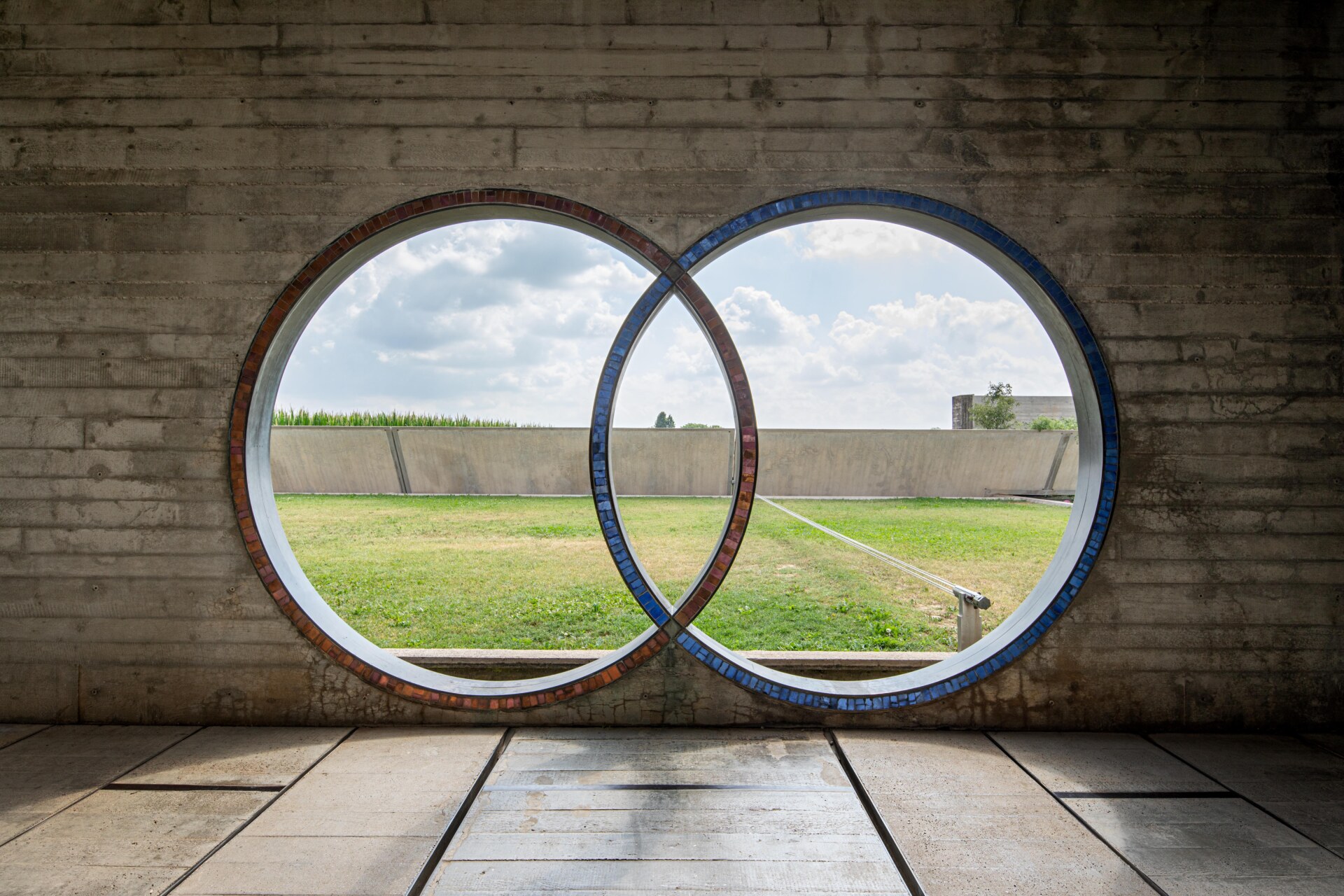 Tomba Brion - Alt Text: Concrete wall with two interlocking circular windows, one edged in red and the other in blue. The view through the windows shows a green grassy field and a cloudy sky, framed by parts of the surrounding structure.