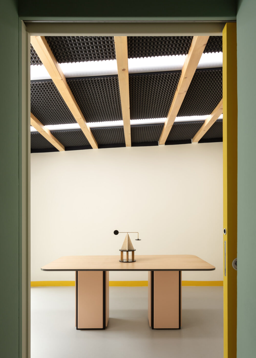 Un ufficio minimalista con un tavolo in legno, due gambe rettangolari e un piccolo elemento decorativo geometrico sul piano superiore. Il soffitto è caratterizzato da travi in legno e pannelli acustici neri. Le pareti sono di colore chiaro con un accento giallo.