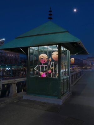 Edicola Radetzky - Two large cartoonish figures are inside a small, illuminated booth at night. One holds a pink guitar and the other holds a microphone. The booth is located on a bridge with city lights and a bright moon in the background.