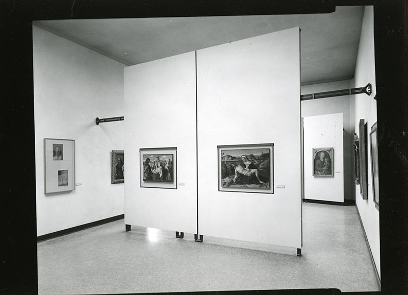 Mostra di una galleria d'arte" - Foto in bianco e nero di una galleria d'arte con diversi dipinti incorniciati su pareti divisorie. La stanza è vuota, con un pavimento lucidato e un soffitto alto. I dipinti variano in dimensioni, raffigurando scene e stili diversi.