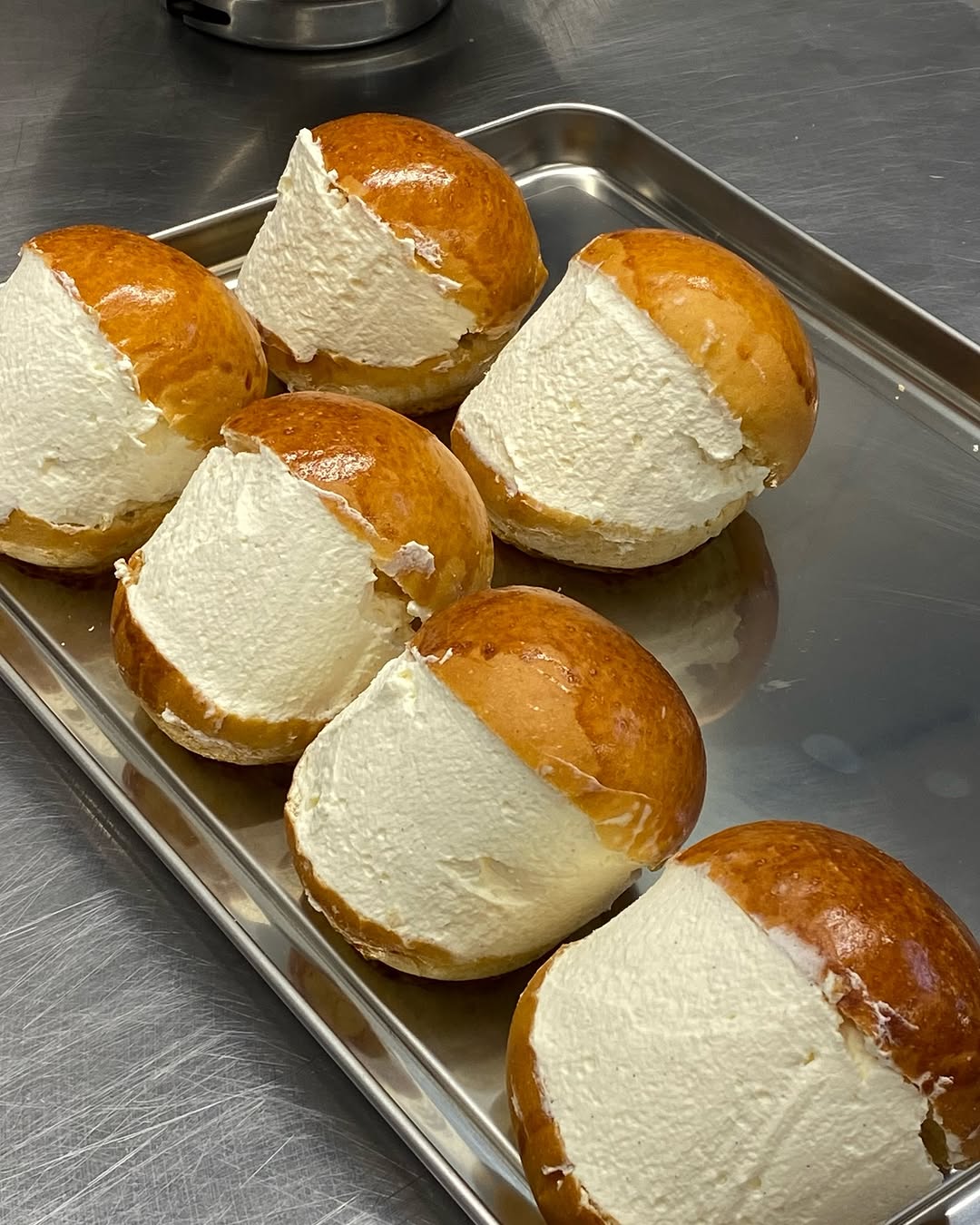Six maritozzi - brioche buns filled generously with white whipped cream, arranged on a rectangular stainless steel tray on a metal countertop.