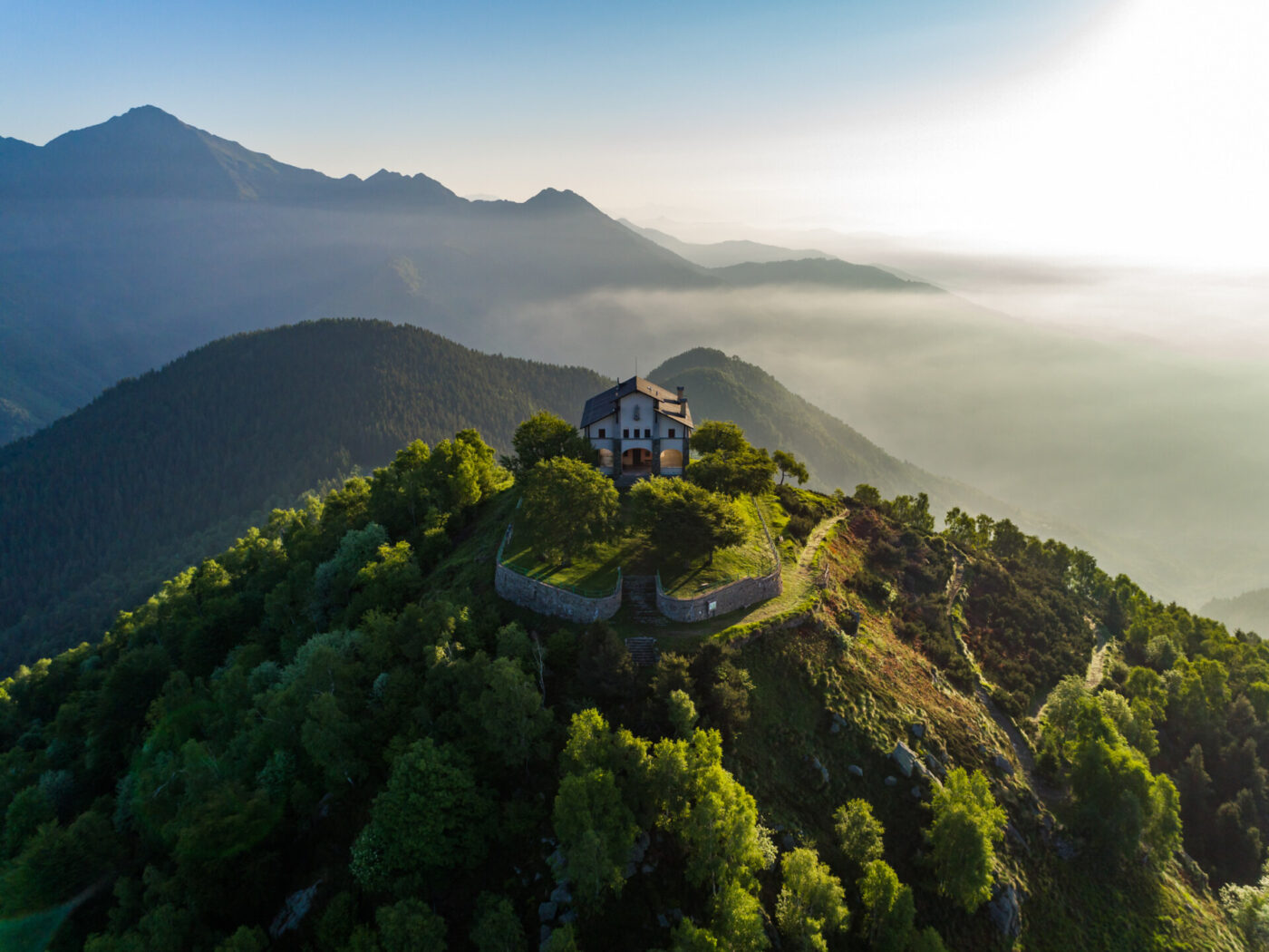 Vista dell'alba dalla cima della collina: una casa in cima alla collina, circondata da alberi e da un muro di pietra, si affaccia sulle montagne nebbiose sotto un cielo azzurro e limpido all'alba.