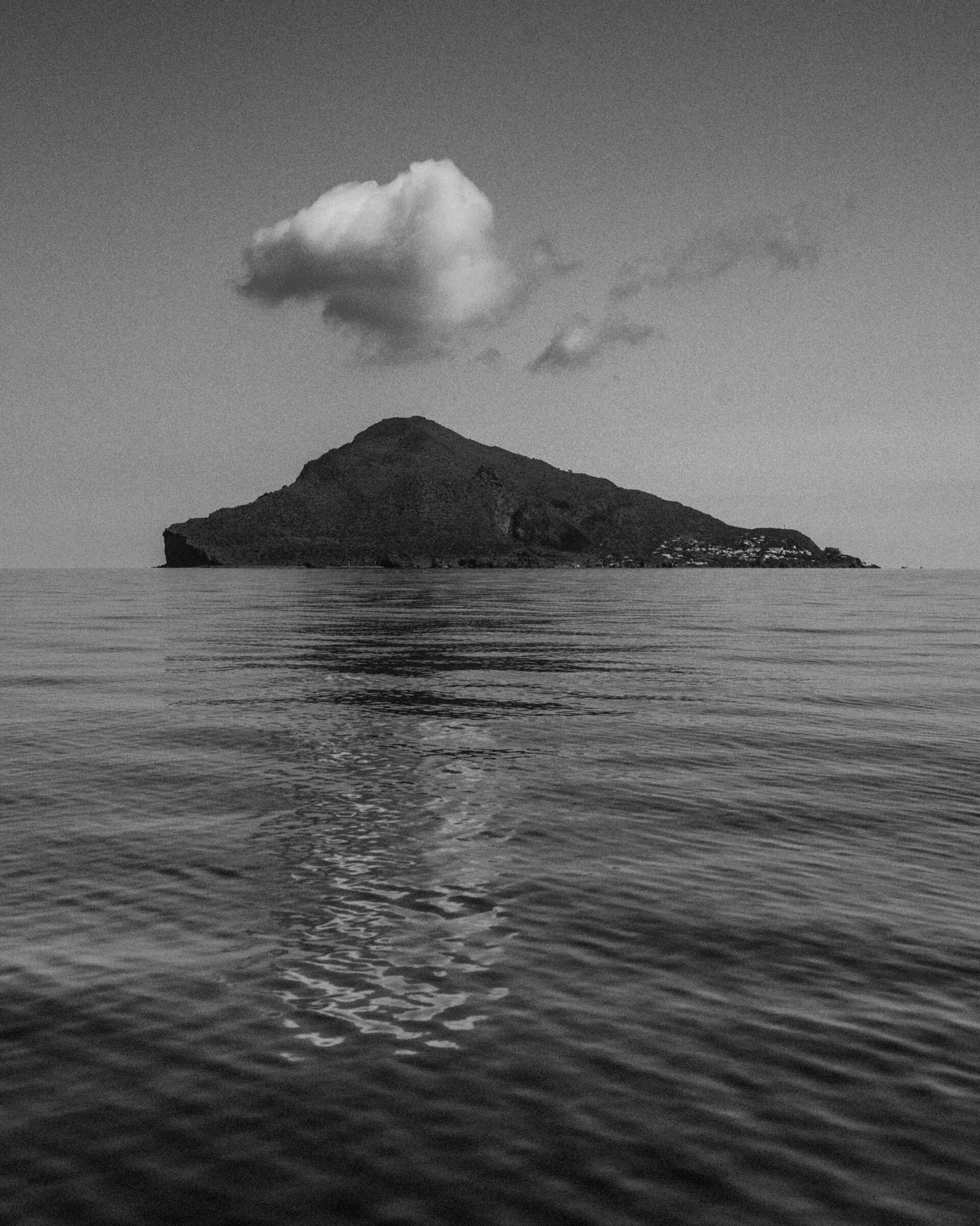 Island Solitude - A black-and-white photo of a solitary island with a single cloud above it, reflected on the calm, rippling surface of the surrounding sea.