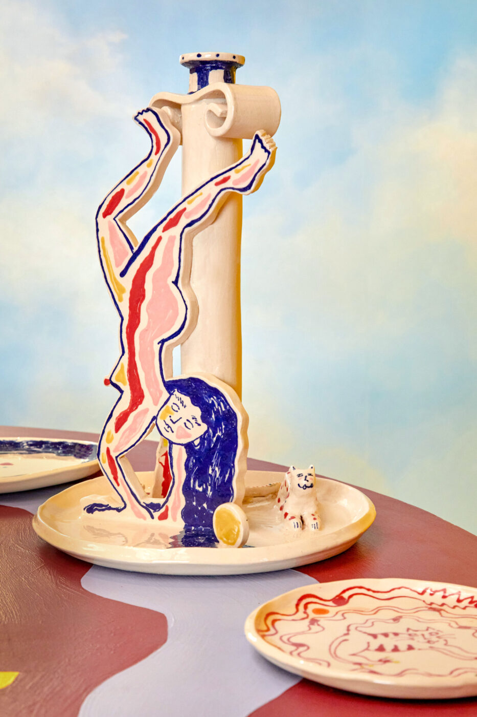 Scultura in ceramica raffigurante una verticale: una colorata scultura in ceramica raffigurante una figura stilizzata e astratta che esegue una verticale su una base rotonda, con una piccola statuetta di un cane accanto e piatti dipinti a mano su un tavolo, il tutto su uno sfondo di cielo nuvoloso e sfumato.