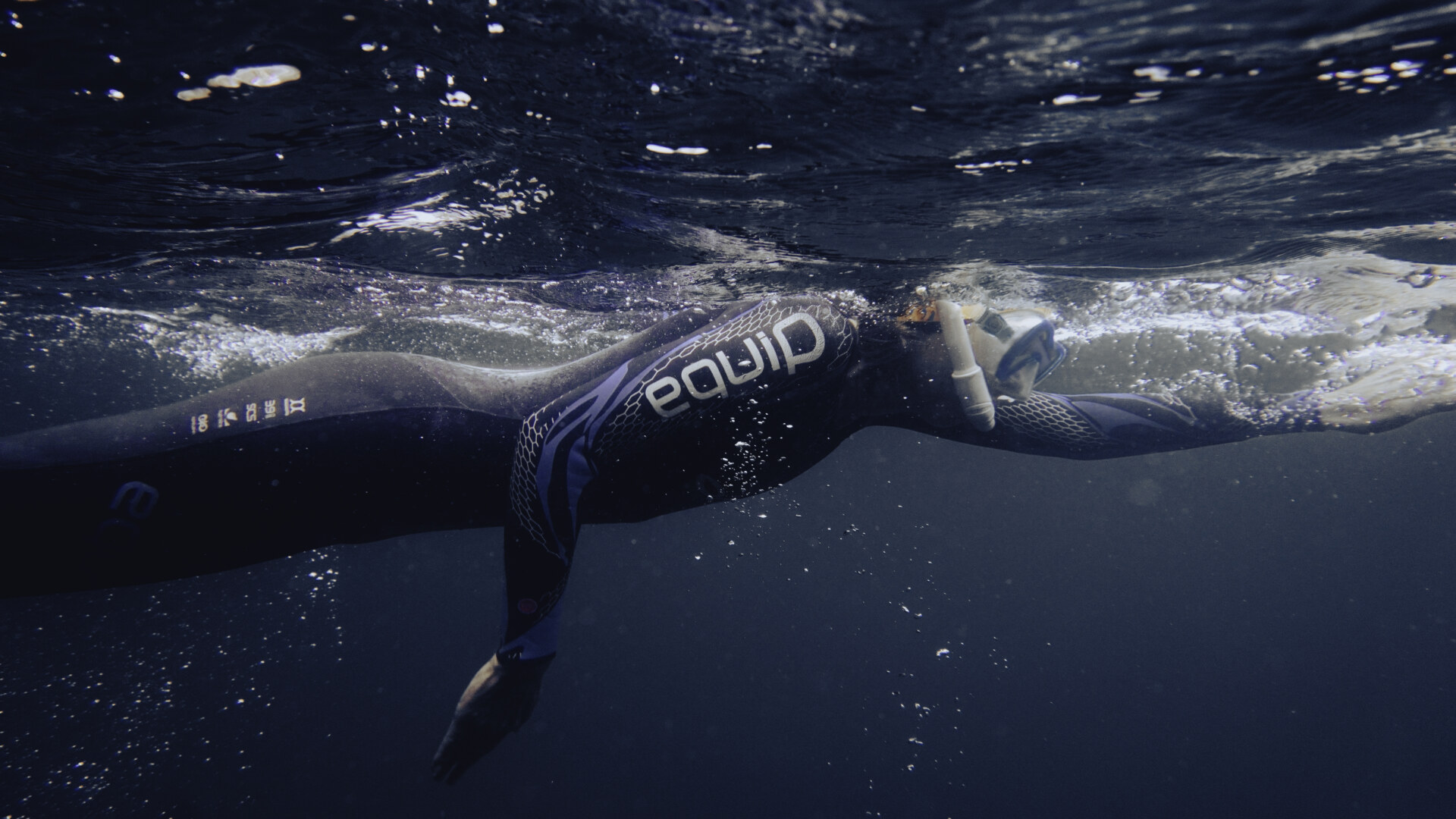 A swimmer wearing a wetsuit and snorkel swims underwater in dark, clear water, reaching forward with one arm extended.