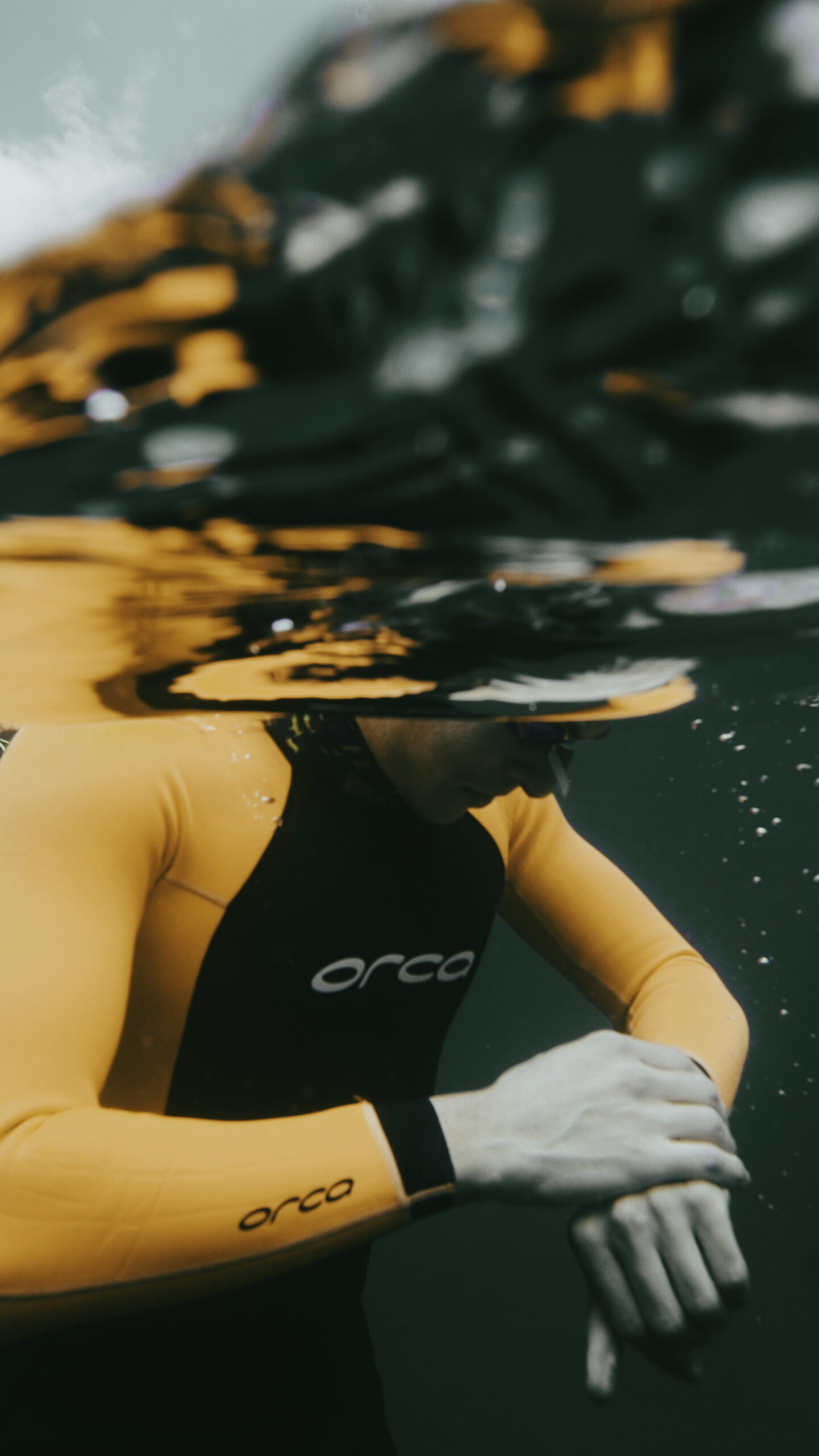 A person wearing a yellow and black wetsuit labeled orca is underwater, looking down at their wrist, possibly checking a watch or device, with water surface reflections above.