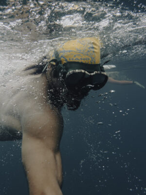 Swimtrekking - A person wearing a yellow swim cap and goggles swims underwater, reaching forward with one arm, surrounded by bubbles in deep blue water.
