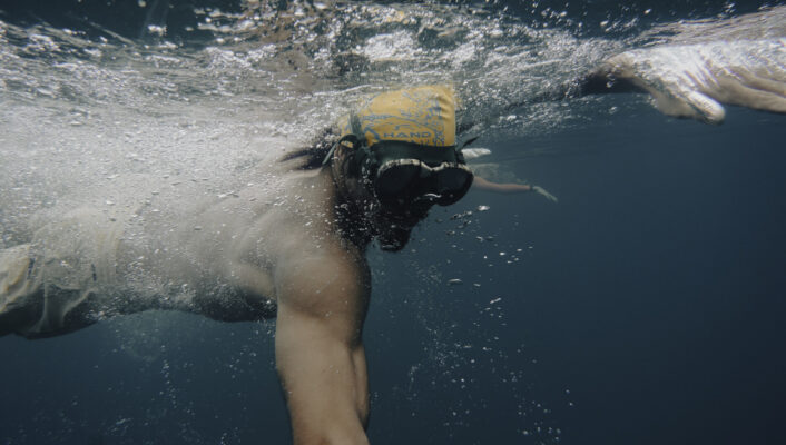 Swimtrekking - A person wearing a yellow swim cap and goggles swims underwater, reaching forward with one arm, surrounded by bubbles in deep blue water.
