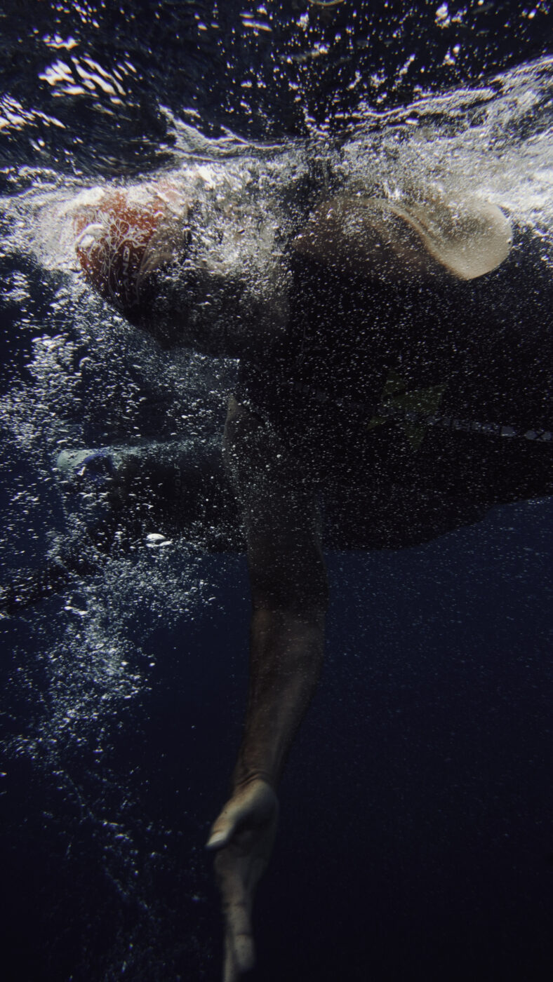Underwater Swimmer - Underwater view of a swimmer in a cap and goggles, captured mid-stroke with one arm extended and bubbles swirling around in dark, deep water.