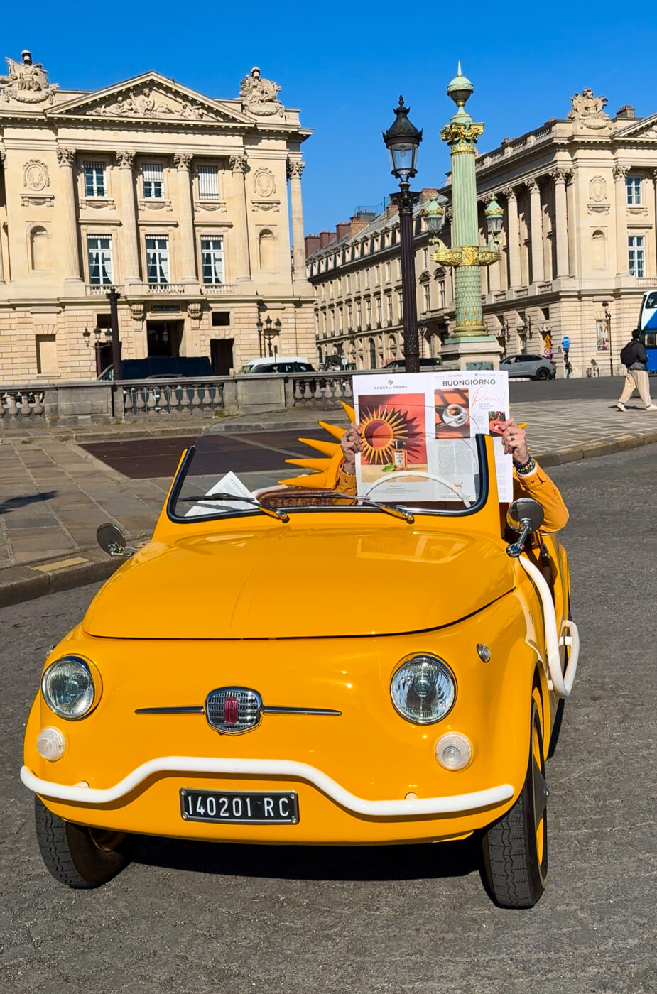 Sunflower Drive - A yellow vintage car is parked on a street in front of historic buildings. The driver, holding a booklet with sunflowers, is partially visible. The surrounding architecture features grand facades and decorative lampposts, under a clear blue sky.