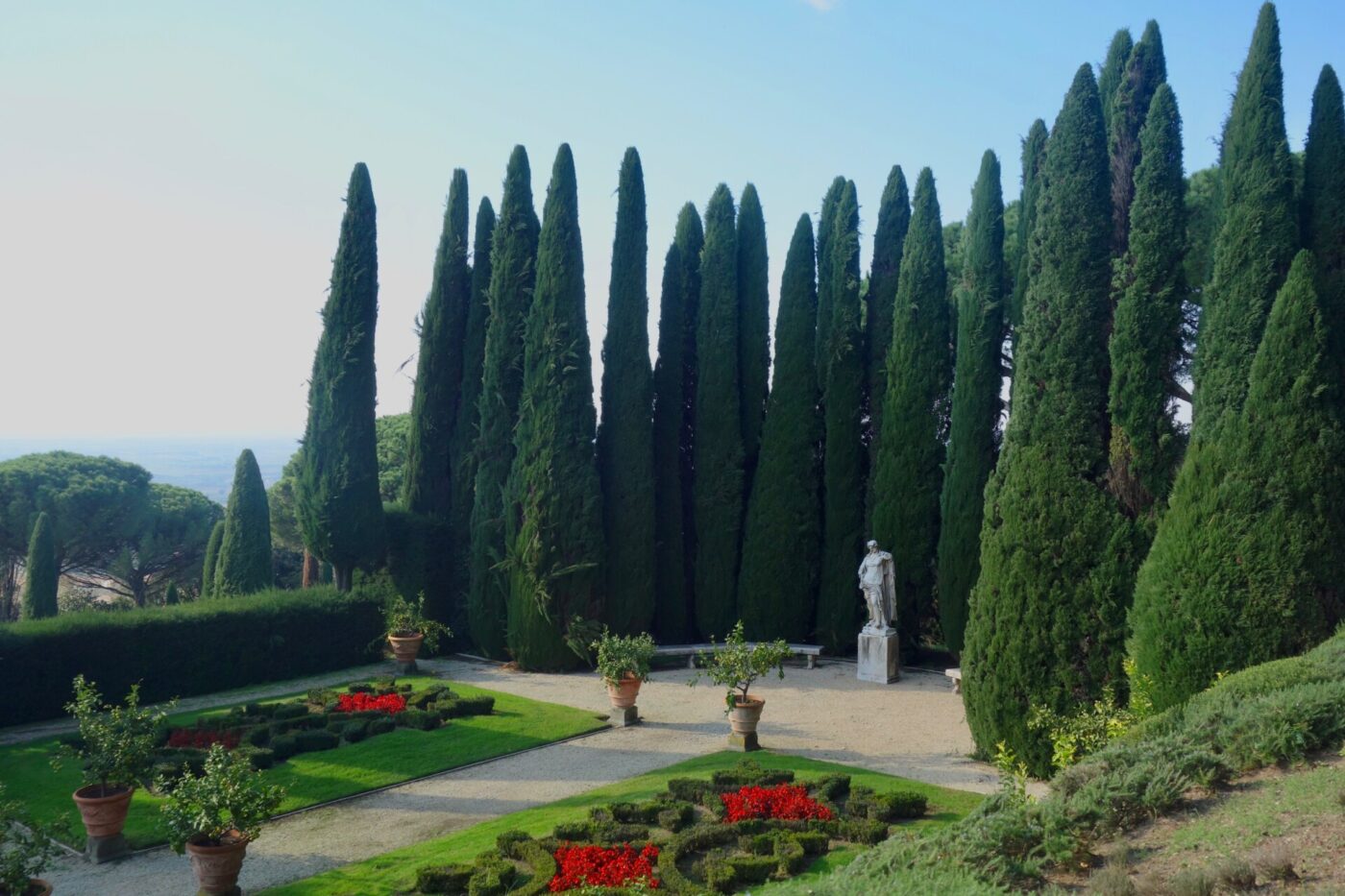 Un giardino formale con siepi curate, aiuole di fiori rossi, piante in vaso e una statua bianca si erge di fronte a un'alta fila di snelli cipressi sotto un cielo azzurro e terso.