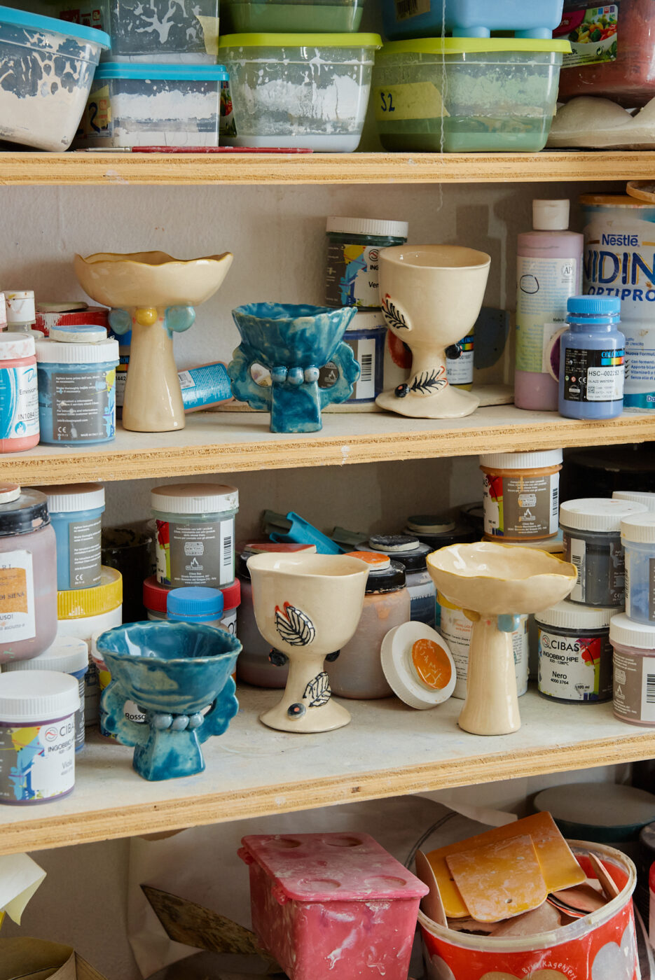 Scaffali pieni di calici e ciotole di ceramica, alcuni smaltati di blu e altri beige con disegni dipinti, circondati da vari contenitori di plastica colorati e barattoli di materiale artistico.