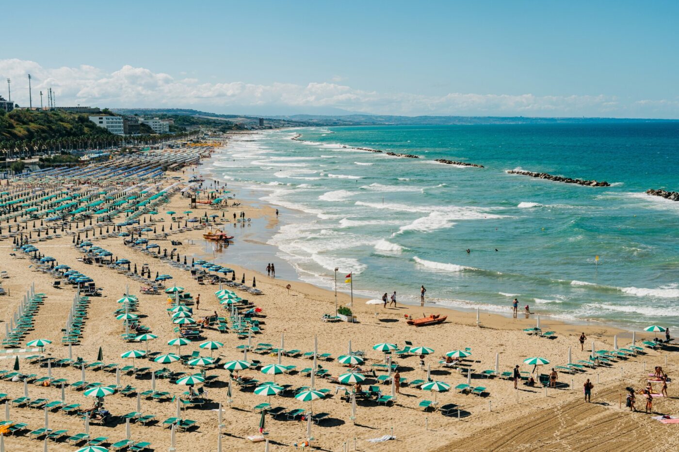 Termoli - Aerial view of a busy beach with rows of green and white umbrellas and sun loungers. People are sunbathing and swimming. The turquoise waves gently wash ashore under a clear blue sky. Buildings and a cliff are visible in the distance.