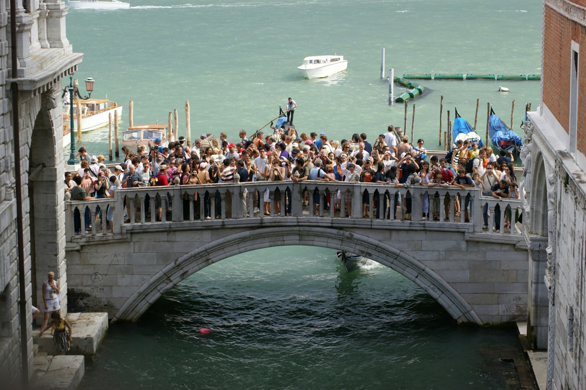 Venice - A large crowd of people stand closely packed on a stone bridge over a canal in Venice, with boats and turquoise water in the background.