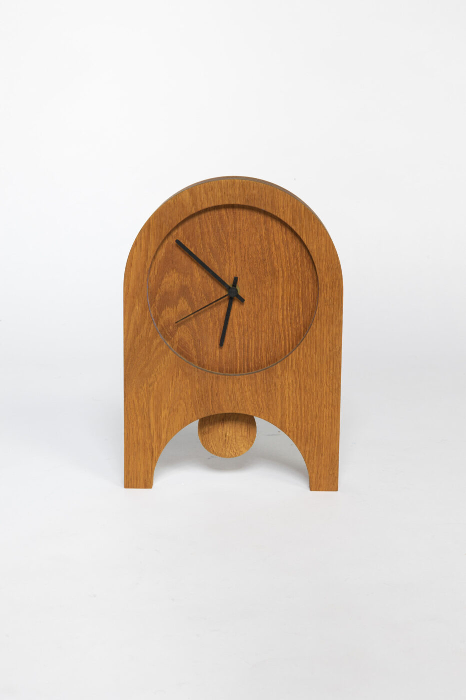 Ophelia Pendulum - A wooden clock with a minimalist design stands on a white background. It has a round face with black hands, and a pendulum hangs below in a matching wood finish.