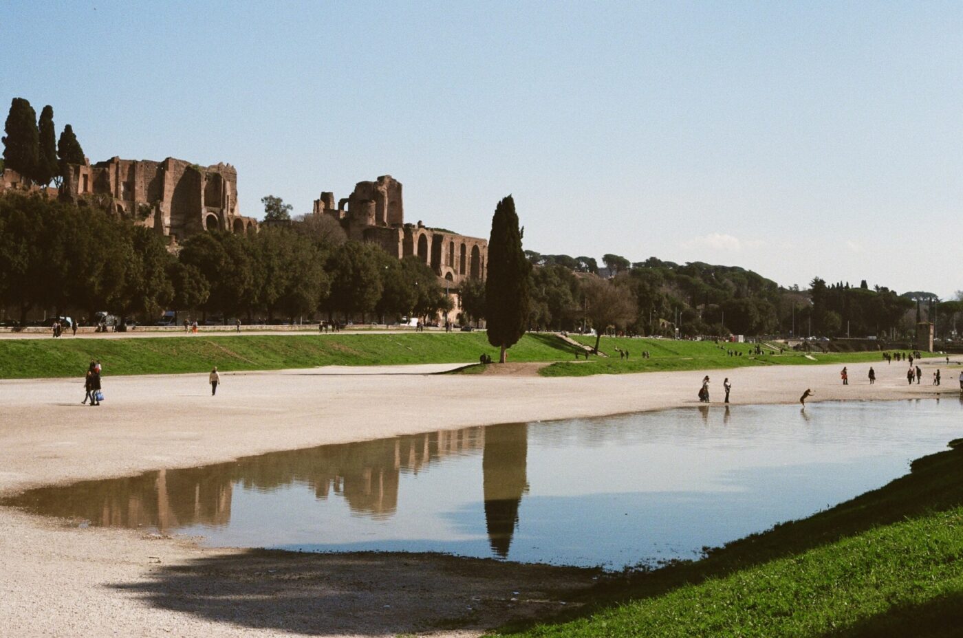 Circus Maximus - People walk and relax around a large, shallow pool at the Circus Maximus in Rome, with ancient ruins and trees in the background under a clear blue sky.