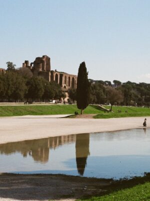 Circus Maximus - People walk and relax around a large, shallow pool at the Circus Maximus in Rome, with ancient ruins and trees in the background under a clear blue sky.