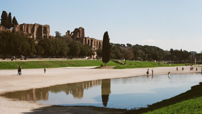 Riflesso al Circo Massimo - L'immagine mostra persone che camminano vicino a una grande pozzanghera poco profonda al Circo Massimo a Roma, con antiche rovine, alberi e un cielo limpido sullo sfondo. Le rovine e gli alberi si riflettono nell'acqua.