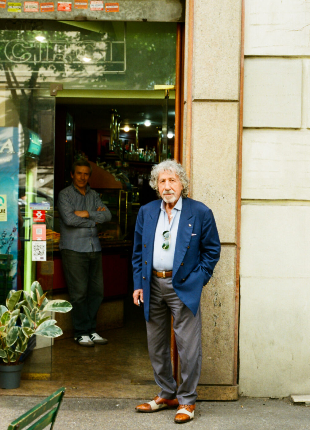 Annotato! Ecco il testo alternativo con il titolo dell'immagine all'inizio, come da tue istruzioni: Titolo dell'immagine: un uomo anziano con i capelli grigi e ricci, in giacca blu, è in piedi fuori da un bar, con le mani in tasca. Un altro uomo è in piedi all'interno, vicino alla porta. All'ingresso ci sono delle piante in vaso; l'interno del bar è illuminato da luci soffuse.