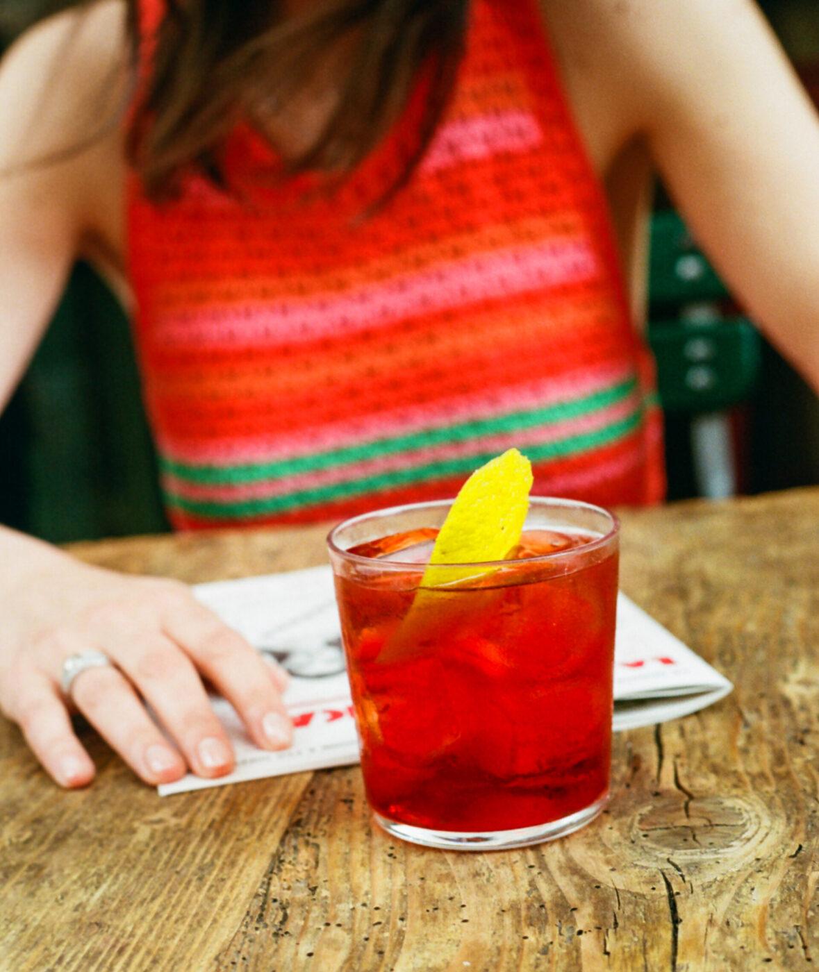 [Nessun titolo dell'immagine presente] - Una persona con una maglietta senza maniche a righe rosse e rosa è seduta a un tavolo di legno con una rivista e un bicchiere di cocktail rosso guarnito con una scorza di limone.