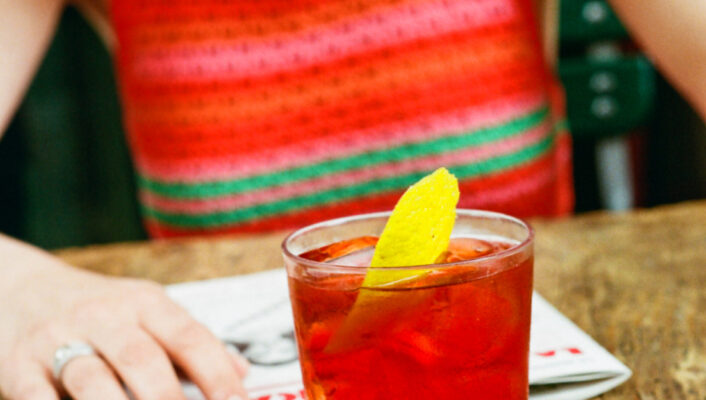 A person in a red and pink striped sleeveless top sits at a wooden table with a magazine and a glass of red cocktail garnished with a lemon twist.