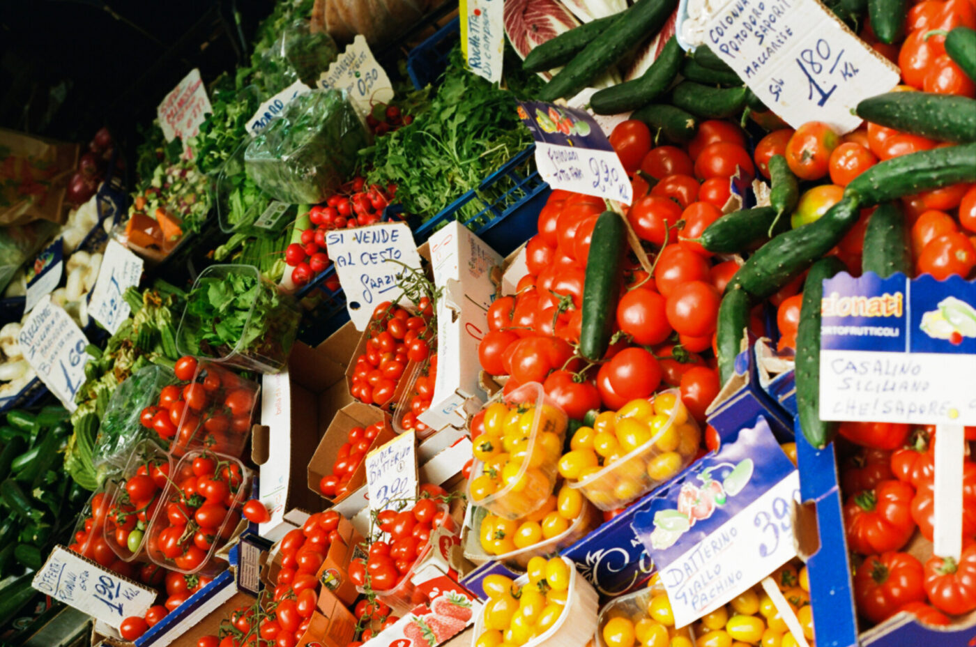Esposizione colorata di verdure fresche al mercato: verdure fresche come pomodori rossi e gialli, cetrioli e verdure a foglia verde sono disposte in scatole e cesti con cartellini dei prezzi scritti a mano in italiano.