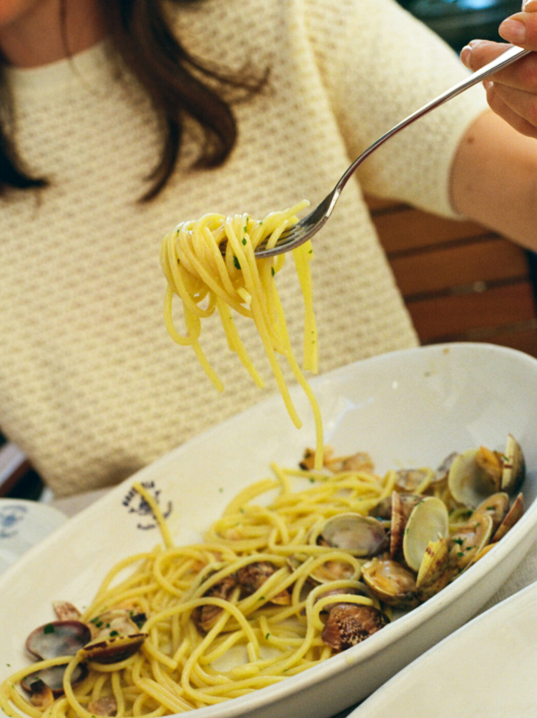 [Nessun titolo dell'immagine] - Una persona con una maglia color crema solleva una forchettata di spaghetti da un grande piatto bianco di pasta con vongole.