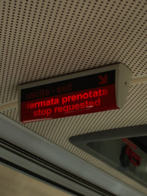 Uscita - exit, fermata prenotata - stop requested - Un cartello elettronico all'interno di un veicolo mostra "uscita - exit" e "fermata prenotata - stop requested" in rosso, indicando che è stata richiesta una fermata. Il cartello è montato su un soffitto forato.