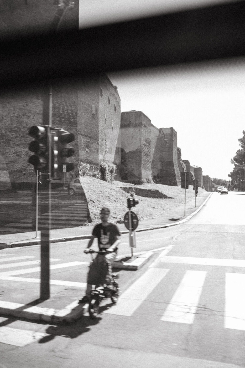 Bicycle Ride Near Stone Walls - A person rides a bicycle across a crosswalk near ancient stone walls, with a traffic light and street signs nearby. The scene is captured in black and white.