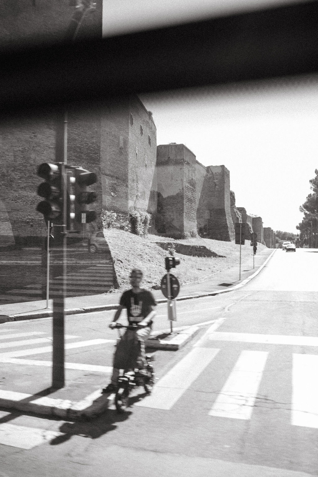 Bicycle Ride Near Stone Walls - A person rides a bicycle across a crosswalk near ancient stone walls, with a traffic light and street signs nearby. The scene is captured in black and white.