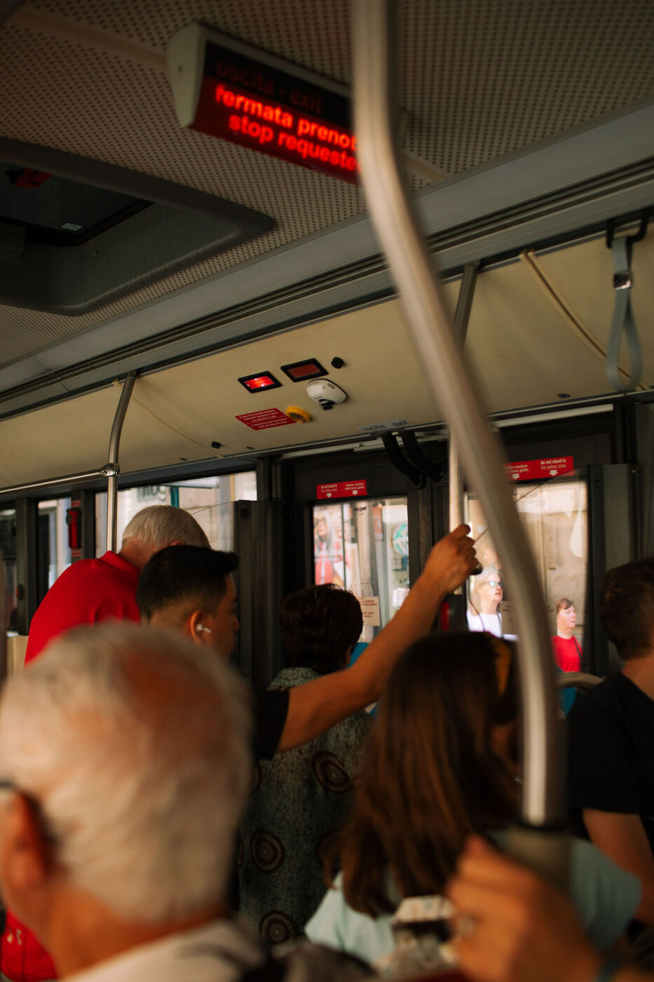 Usicta Exit Fermata Prenotata Stop Requested - A group of people stand and hold onto poles inside a crowded bus. An electronic sign above displays “uscita exit fermata prenotata stop requested” in red letters. Sunlight streams through the windows, illuminating the passengers.