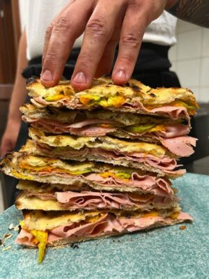Pila alta di panini - Una mano tiene ferma una pila alta di panini tostati, farciti con affettati sottili, verdure e formaggio fuso, il tutto sistemato su un piatto blu in una cucina.