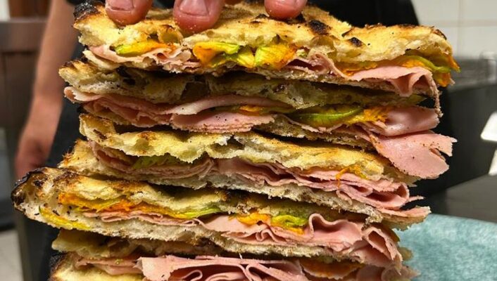 Pila alta di panini - Una mano tiene ferma una pila alta di panini tostati, farciti con affettati sottili, verdure e formaggio fuso, il tutto sistemato su un piatto blu in una cucina.