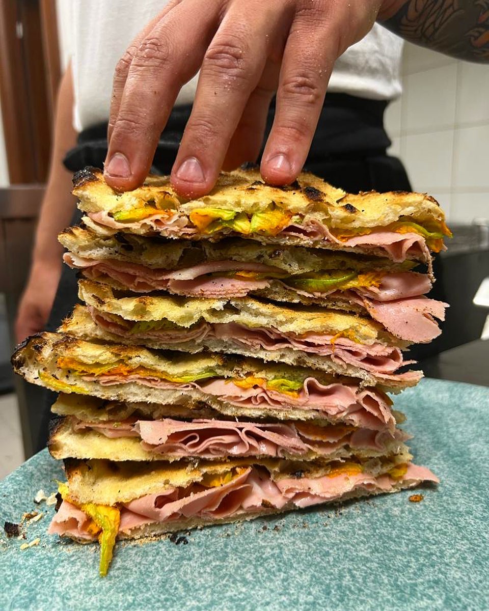 Pila alta di panini - Una mano tiene ferma una pila alta di panini tostati, farciti con affettati sottili, verdure e formaggio fuso, il tutto sistemato su un piatto blu in una cucina.