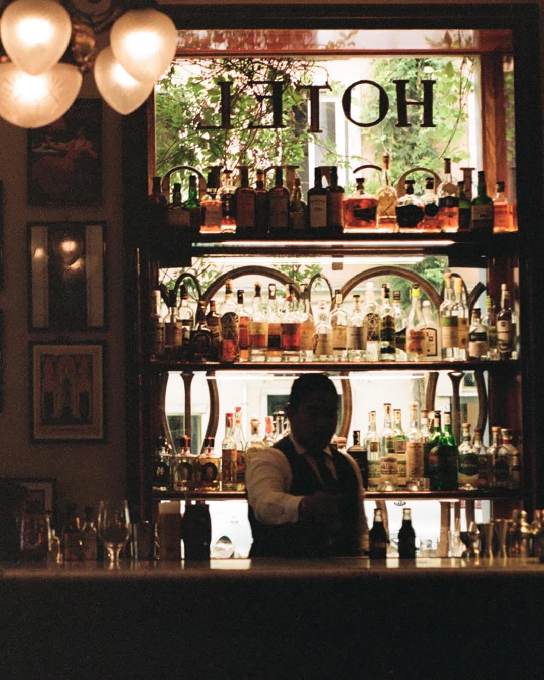 A bartender stands behind a dimly lit bar, surrounded by shelves filled with bottles, with sunlight streaming through a window that has HOTEL written on it in reverse.