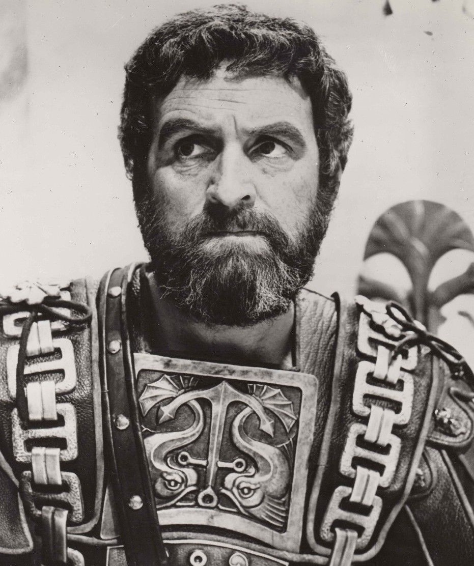Andrew Keir in Cleopatra - A man with dark curly hair and a beard wears ornate, ancient-style armor with patterned designs, looking slightly to the side with a serious expression. The image is in black and white.