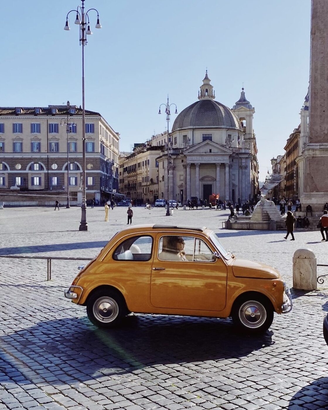 Piccola auto gialla in una piazza europea - Una piccola auto gialla d'epoca attraversa una piazza acciottolata in una città europea, con edifici storici, cupole e persone che camminano sullo sfondo sotto un cielo azzurro e limpido.