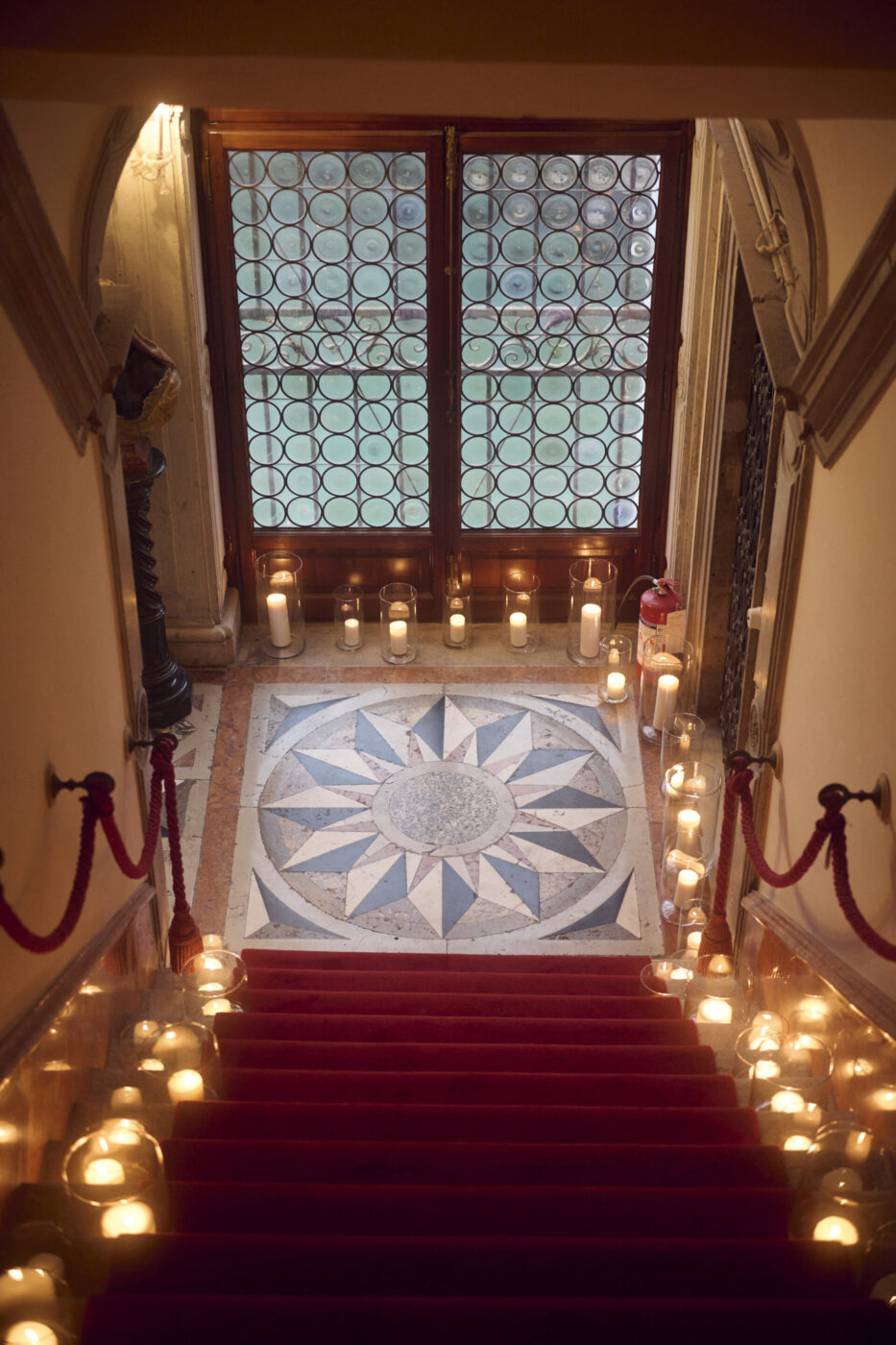Una grande scalinata con tappeto rosso è fiancheggiata da candele accese in portacandele di vetro e conduce a un pavimento a mosaico con motivo a stelle e grandi porte di legno con pannelli di vetro circolari.