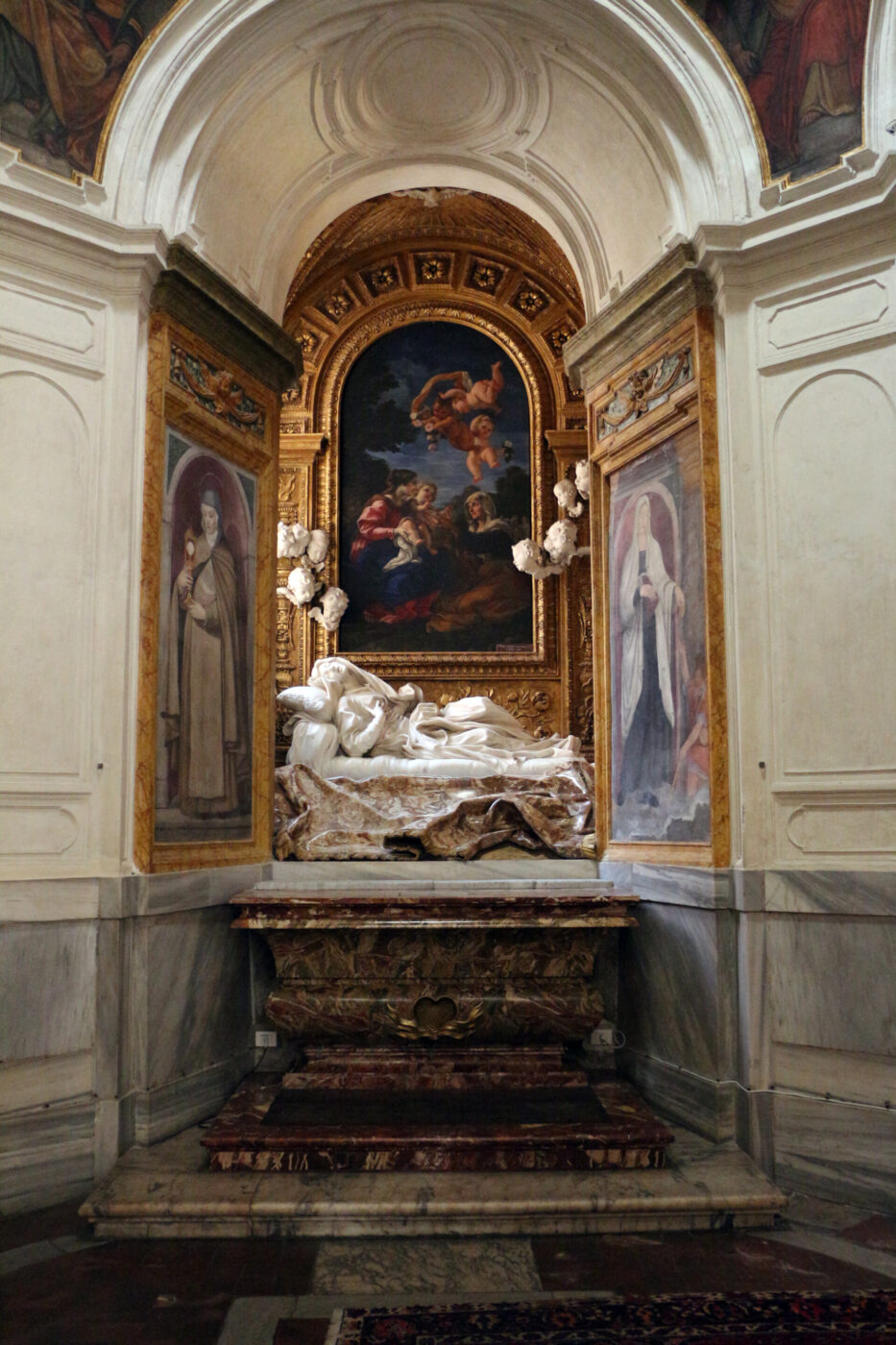 [Nessun titolo dell'immagine fornito] - Una cappella decorata presenta una scultura in marmo di una figura sdraiata, affiancata da ritratti dipinti di santi, sotto un arco dorato con un dipinto religioso sopra; la scena è ambientata in un'alcova riccamente decorata e scarsamente illuminata.