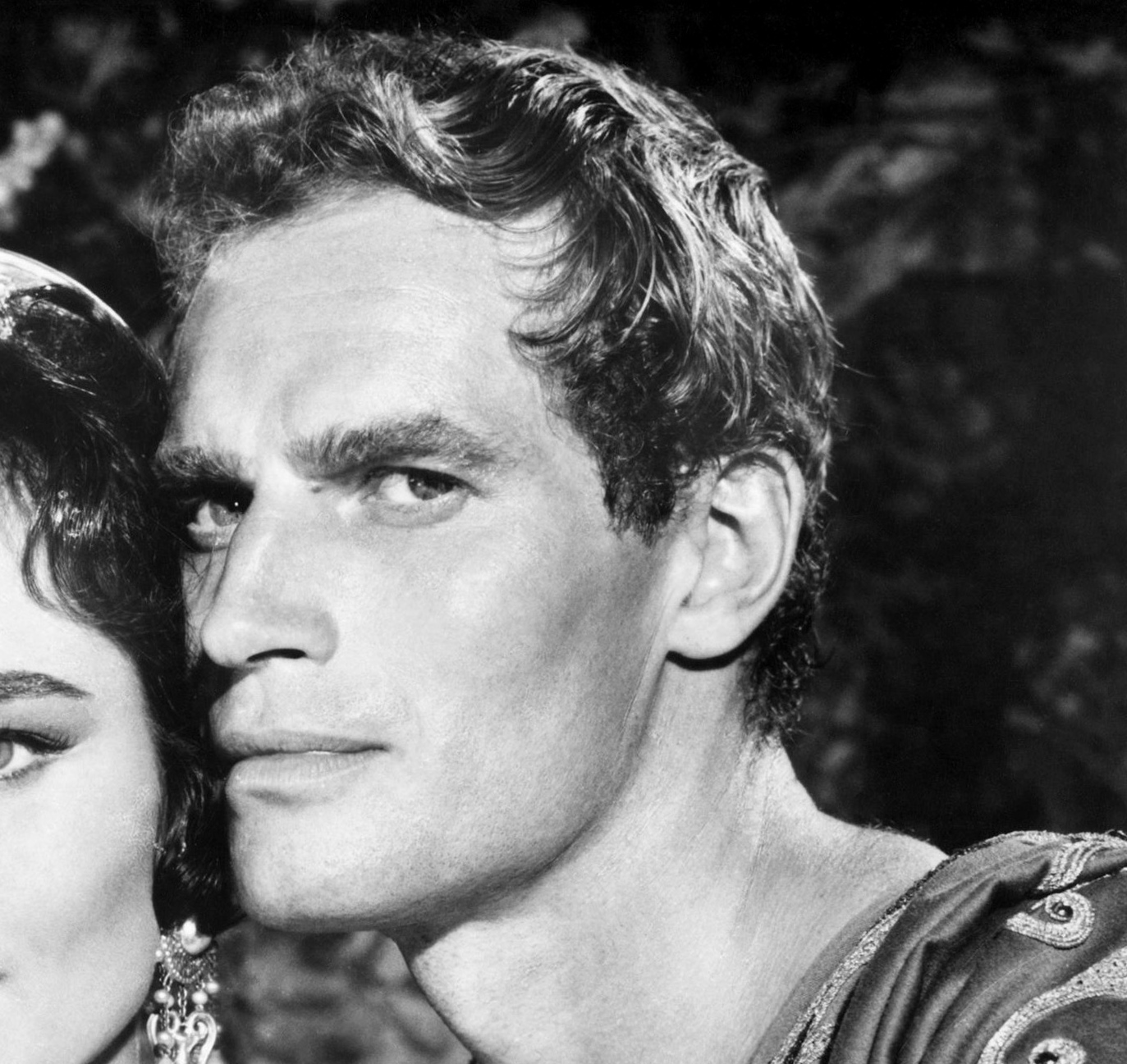 A black-and-white close-up photo of Charlton Heston for Ben-Hur with short, curly hair and strong facial features, gazing seriously at the camera. A woman’s face is partially visible next to him on the left side of the image.