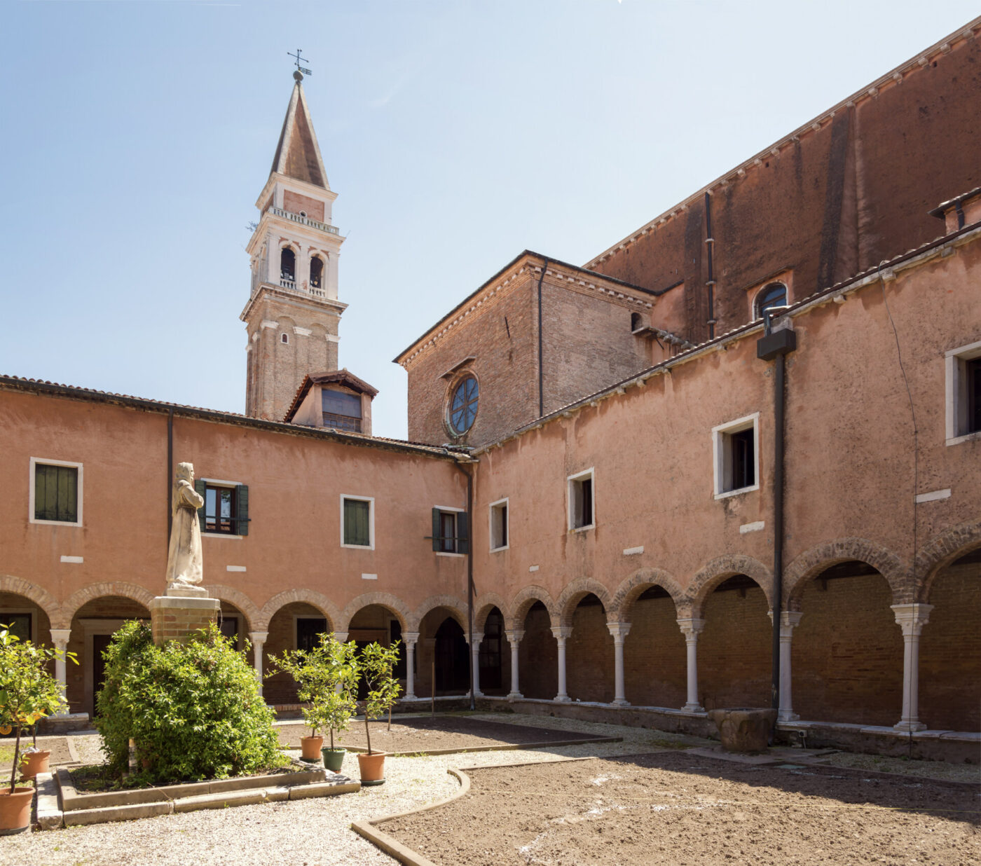 [Titolo dell'immagine] - Un cortile tranquillo con una statua centrale, circondato da passaggi ad arco e piante in vaso, e un alto campanile che si erge dietro l'edificio storico color marrone-rossastro sotto un cielo limpido.