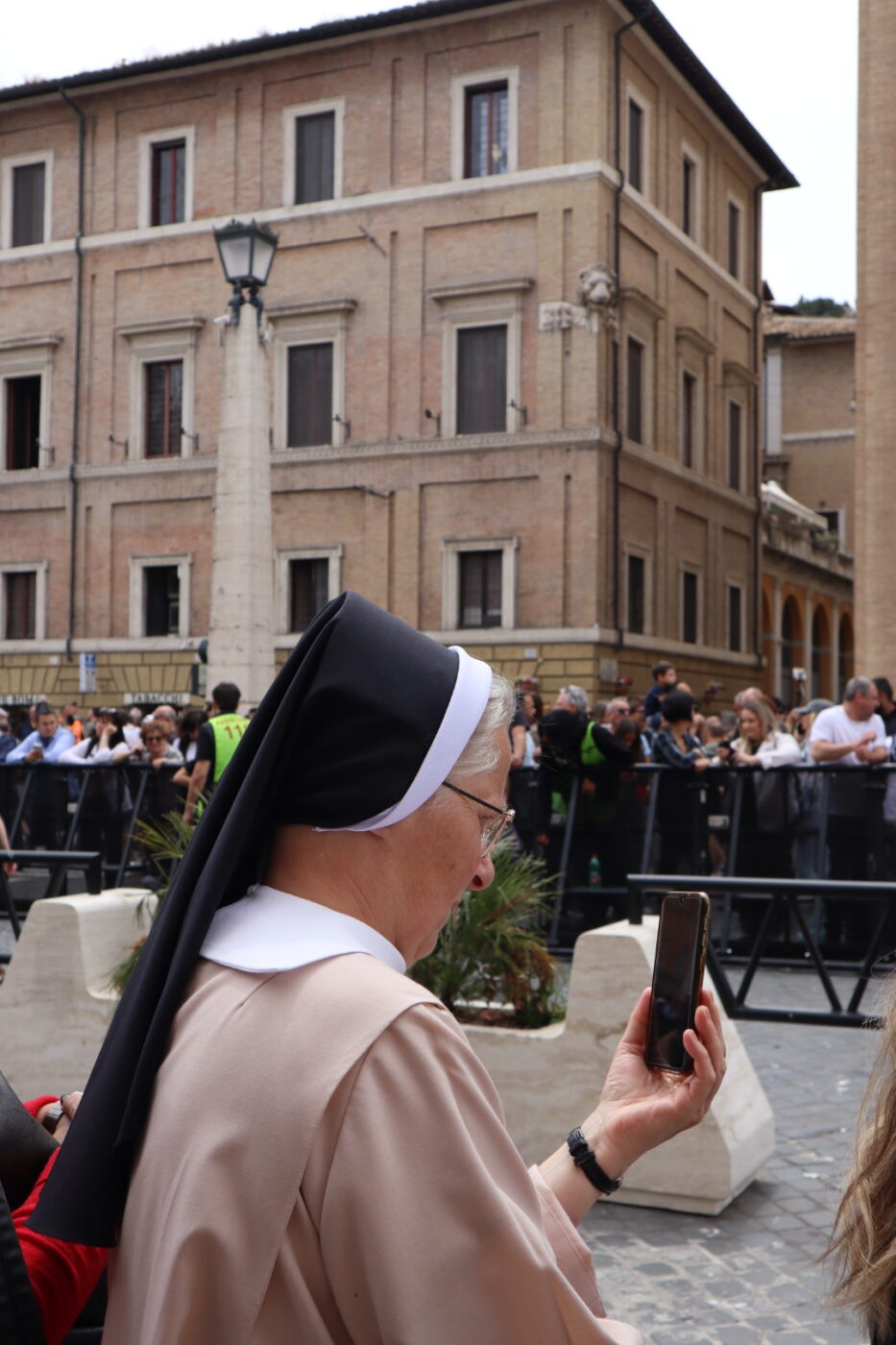 Nun photographing crowd - A nun wearing a black and white habit holds up a smartphone, taking a photo or video in a crowded outdoor area with people and historic buildings in the background.