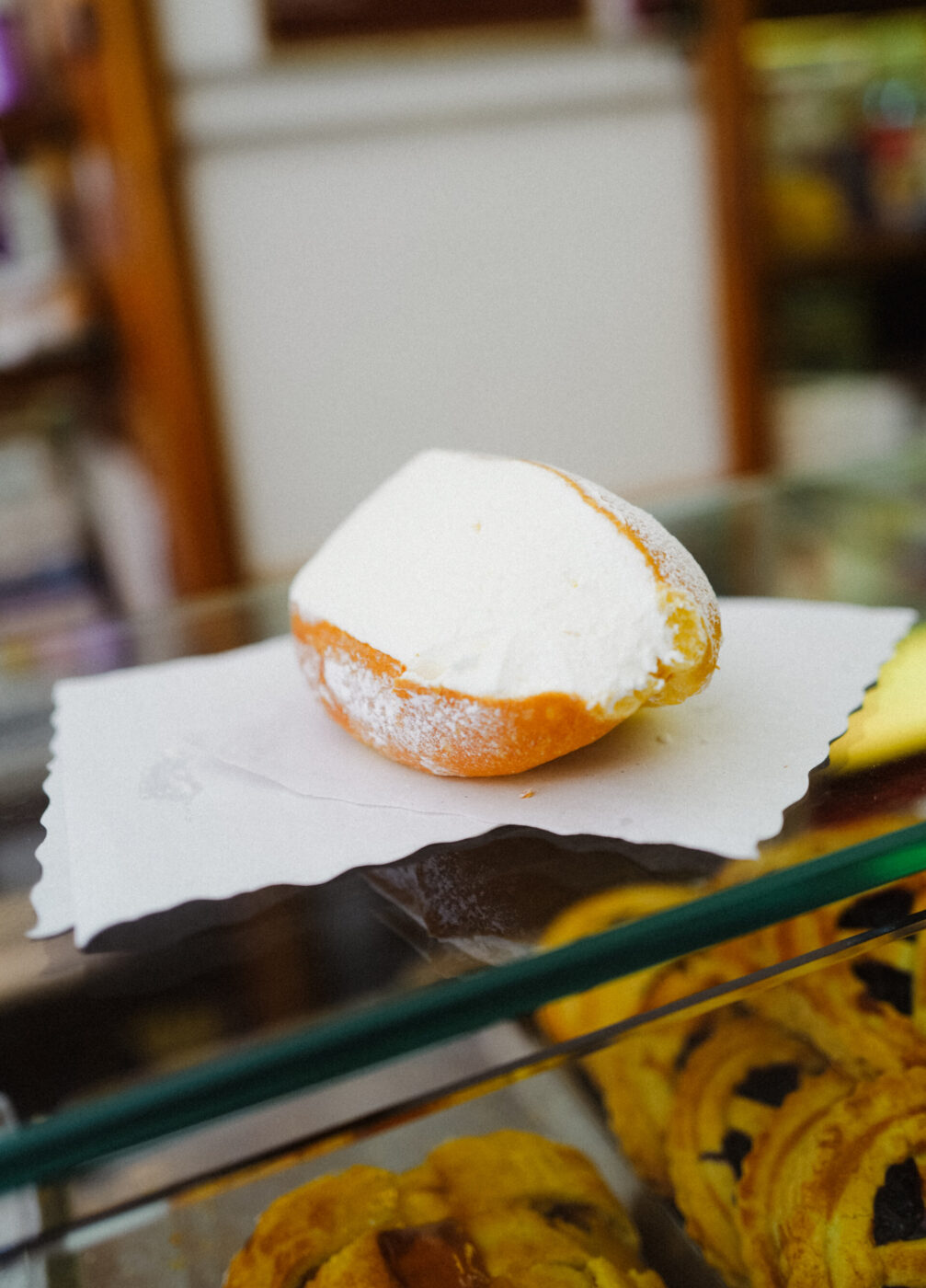 Maritozzo con ricotta - Un dolce siciliano ripieno di ricotta, il maritozzo, è adagiato su un tovagliolo bianco quadrato, sopra una vetrina di vetro in una pasticceria. Il dolce è spolverato di zucchero a velo.