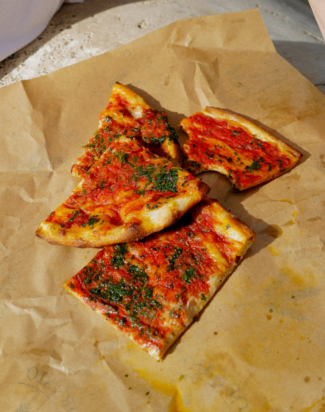 Quattro fette di pizza sottile - Quattro fette di pizza sottile con salsa di pomodoro ed erbe aromatiche vengono disposte su carta da forno marrone, su una superficie chiara e ruvida.
