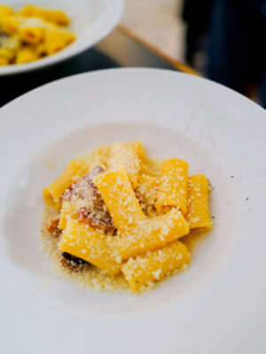 Carbonara: A white plate filled with rigatoni pasta, topped with grated cheese and bits of bacon, sits on a dark table. Another plate of pasta is blurred in the background.